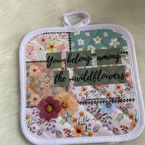 May include: White quilted pot holder with a floral print and the text "You belong among the wildflowers".