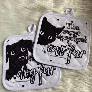 May include: Two white quilted pot holders with black cat and dog silhouettes. The cat pot holder says "The secret ingredient is cat fur." The dog pot holder says "The secret ingredient is dog fur."