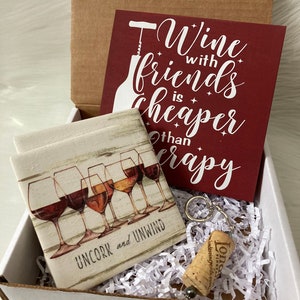 May include: A gift box containing a coaster set with wine glasses, a keychain with a cork, and a burgundy sign that reads "Wine with friends is cheaper than therapy."
