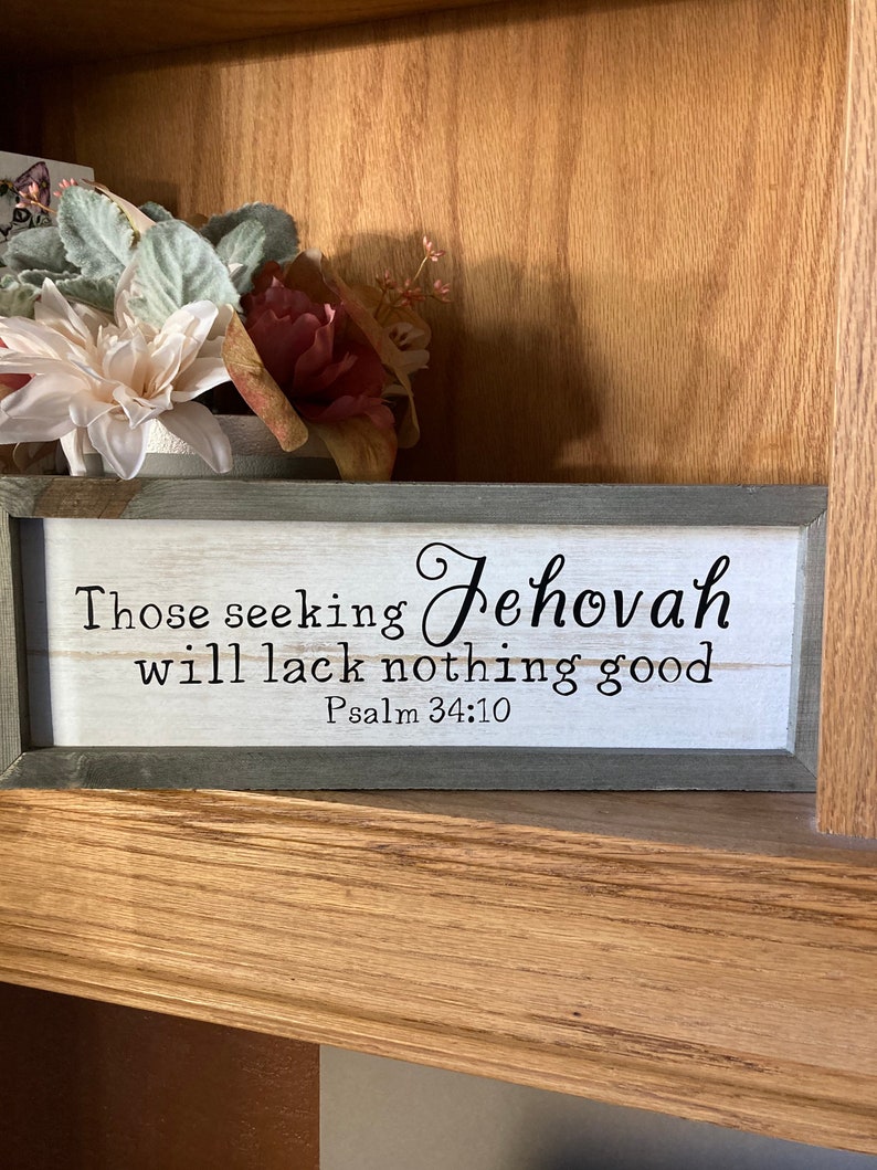 May include: A wooden sign with a white background and black lettering. The sign reads "Those seeking Jehovah will lack nothing good" and "Psalm 34:10". The sign is framed in a gray wood frame.