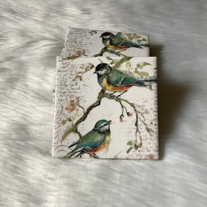 May include: Two decorative tiles with a white background and a vintage floral design. The tiles feature bluebirds perched on branches with red berries. The tiles have a distressed look with faded text and a white background.