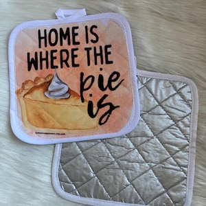 May include: Two square quilted pot holders. One features a watercolor image of a pie slice with whipped cream and the text "HOME IS WHERE THE pie is". The other is silver with a quilted pattern and white trim.