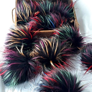 May include: A collection of colorful faux fur pom poms in various shades of black, red, green, and gold. The pom poms are arranged in a wooden box.