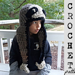 May include: A crocheted raccoon hat with a black and brown striped scarf. The hat has a black nose and eyes and a white face.