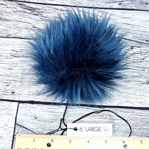 Puede incluir: Un gran pomp&oacute;n de piel sint&eacute;tica azul marino y esponjoso. El pomp&oacute;n mide aproximadamente 15 cm de di&aacute;metro. Una etiqueta adjunta al pomp&oacute;n dice "6" LARGE". El pomp&oacute;n est&aacute; sobre una superficie de madera blanca y azul claro.