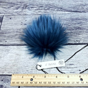 Puede incluir: Un pomp&oacute;n de piel sint&eacute;tica azul oscuro con una etiqueta que dice "4" SMALL". El pomp&oacute;n es redondo y tiene fibras largas y suaves. Una regla debajo muestra las medidas en cent&iacute;metros. El pomp&oacute;n mide unos 10 cm de di&aacute;metro.