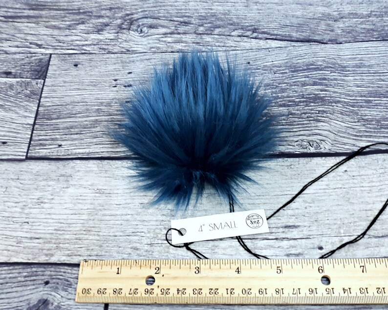 Puede incluir: Un pomp&oacute;n de piel sint&eacute;tica azul oscuro con una etiqueta que dice "4" SMALL". El pomp&oacute;n es redondo y tiene fibras largas y suaves. Una regla debajo muestra las medidas en cent&iacute;metros. El pomp&oacute;n mide unos 10 cm de di&aacute;metro.