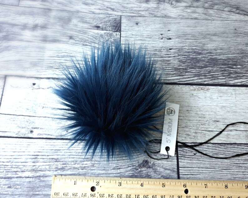 Puede incluir: Un pomp&oacute;n de piel sint&eacute;tica azul marino, de aproximadamente 13 cm de di&aacute;metro, con una etiqueta que dice "5" MEDIUM". El pomp&oacute;n es redondo y esponjoso, con fibras largas y puntiagudas. El fondo es una superficie de madera clara.