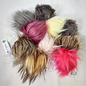 May include: A collection of fluffy faux fur pom poms in various colors, including gray, pink, yellow, and brown. Each pom pom has a tag that reads "4 SMALL". The pom poms are arranged on a white surface.