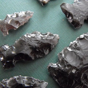 May include: Several dark obsidian arrowheads are arranged on a green surface. The arrowheads have sharp edges and are shaped with a triangular point and a notched base. The obsidian has a glossy, reflective surface.