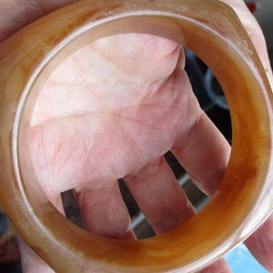 K&ouml;nnte beinhalten: Ein quadratisch geformtes, bernsteinfarbenes Armreif-Armband. Das Armband hat eine glatte, polierte Oberfl&auml;che mit Farbvariationen, die von hellem Beige bis zu einem tieferen Braun reichen. Der Armreif wird in einer Hand gehalten und zeigt seine warmen T&ouml;ne und sein einzigartiges Design.