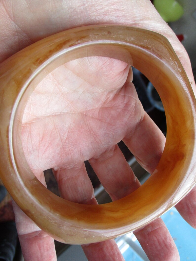 K&ouml;nnte beinhalten: Ein quadratisch geformtes, bernsteinfarbenes Armreif-Armband. Das Armband hat eine glatte, polierte Oberfl&auml;che mit Farbvariationen, die von hellem Beige bis zu einem tieferen Braun reichen. Der Armreif wird in einer Hand gehalten und zeigt seine warmen T&ouml;ne und sein einzigartiges Design.
