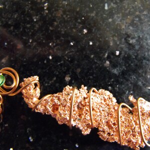 May include: A handcrafted copper necklace featuring a textured, irregular copper pendant. The pendant is wrapped with copper wire and accented with a small, round, green gemstone. The necklace has a delicate copper chain.