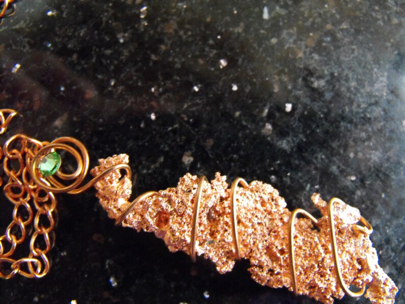 May include: A handcrafted copper necklace featuring a textured, irregular copper pendant. The pendant is wrapped with copper wire and accented with a small, round, green gemstone. The necklace has a delicate copper chain.