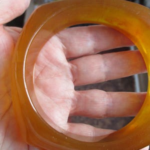 K&ouml;nnte beinhalten: Ein durchscheinendes, bernsteinfarbenes Armreif-Armband mit quadratischer Form. Das Armband besteht aus einem glatten, gl&auml;nzenden Material und hat einen warmen, honigartigen Farbton. Das Licht scheint durch das Armband und hebt seine Farbe und Form hervor.