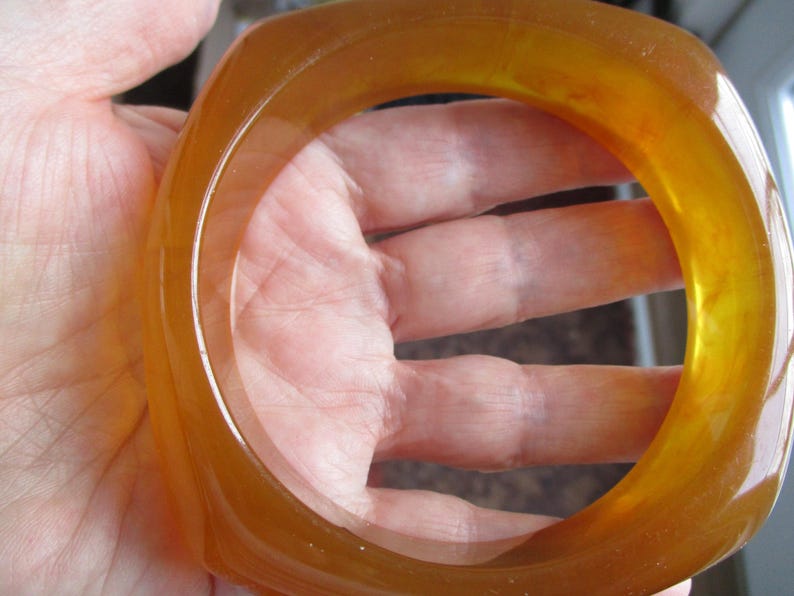 K&ouml;nnte beinhalten: Ein durchscheinendes, bernsteinfarbenes Armreif-Armband mit quadratischer Form. Das Armband besteht aus einem glatten, gl&auml;nzenden Material und hat einen warmen, honigartigen Farbton. Das Licht scheint durch das Armband und hebt seine Farbe und Form hervor.