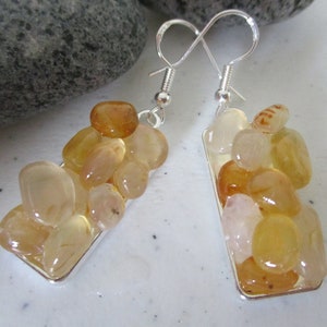 May include: A pair of dangle earrings featuring a cluster of polished, amber-colored stones set in a rectangular silver frame. The stones vary in shade from pale yellow to deep orange, creating a natural, earthy aesthetic. The earrings have silver hooks.