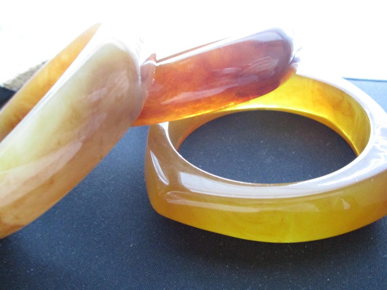 K&ouml;nnte beinhalten: Drei armb&auml;nder in bernsteinfarben. Die Armb&auml;nder haben eine glatte, gl&auml;nzende Oberfl&auml;che und variieren in den Farbt&ouml;nen von hellgelb bis tiefbraun. Die Armb&auml;nder bestehen aus einem durchscheinenden Material, durch das Licht hindurchscheinen kann. Die Armb&auml;nder sind ein Modeaccessoire.