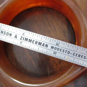K&ouml;nnte beinhalten: Ein bernsteinfarbenes Armreif-Armband mit quadratischem Profil. Ein Lineal liegt &uuml;ber dem Armband, mit dem Text "BENSON & ZIMMERMAN MODESTO-CERES-NEWMAN" sichtbar. Das Armband hat einen Durchmesser von etwa 5 cm.