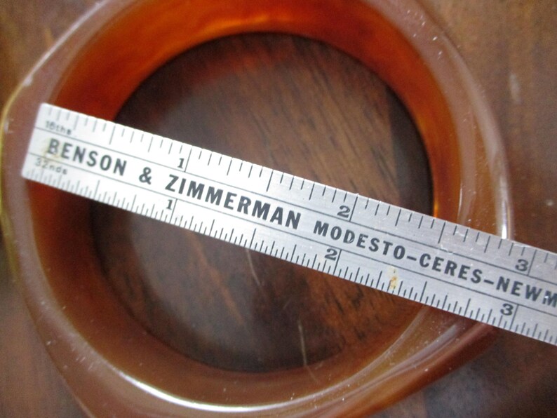 K&ouml;nnte beinhalten: Ein bernsteinfarbenes Armreif-Armband mit quadratischem Profil. Ein Lineal liegt &uuml;ber dem Armband, mit dem Text "BENSON & ZIMMERMAN MODESTO-CERES-NEWMAN" sichtbar. Das Armband hat einen Durchmesser von etwa 5 cm.