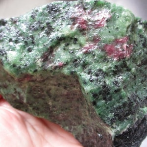May include: A rough-cut gemstone featuring a mix of green, black, and red hues. The stone has a textured surface with dark spots and patches of deep red. The overall appearance is natural and unpolished.