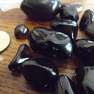 May include: A collection of polished black obsidian stones, varying in size and shape, are scattered on a wooden surface. The stones have a glossy, reflective surface. A US quarter coin is visible, providing a size reference.