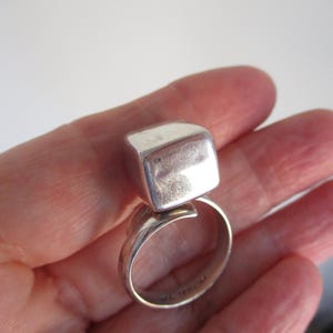 May include: A silver ring with a large, square-shaped top. The ring band curves around the finger, and the square top is slightly rounded. The ring is held in a hand against a white background. The ring is a piece of jewelry.