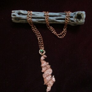 May include: A copper-colored necklace with a unique, textured pendant. The pendant is a freeform shape with a spiral design. The necklace chain is a delicate link style. A small green stone is set in the chain near the pendant. The necklace is displayed on a piece of wood.