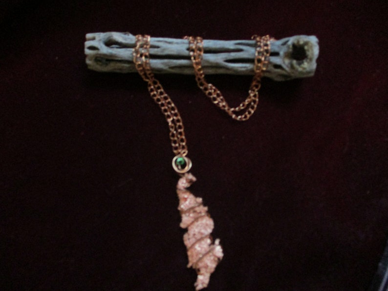 May include: A copper-colored necklace with a unique, textured pendant. The pendant is a freeform shape with a spiral design. The necklace chain is a delicate link style. A small green stone is set in the chain near the pendant. The necklace is displayed on a piece of wood.