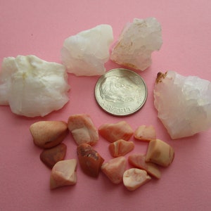 May include: A collection of raw, white quartz crystals and small, polished pink stones are arranged on a pink surface. A US dime is included for size comparison. The quartz pieces have a rough, natural texture.