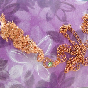 May include: A copper-colored necklace with a pendant featuring a textured, organic design. The pendant is accented with a spiral wire and a small green gemstone. The chain is made of matching copper-colored links.