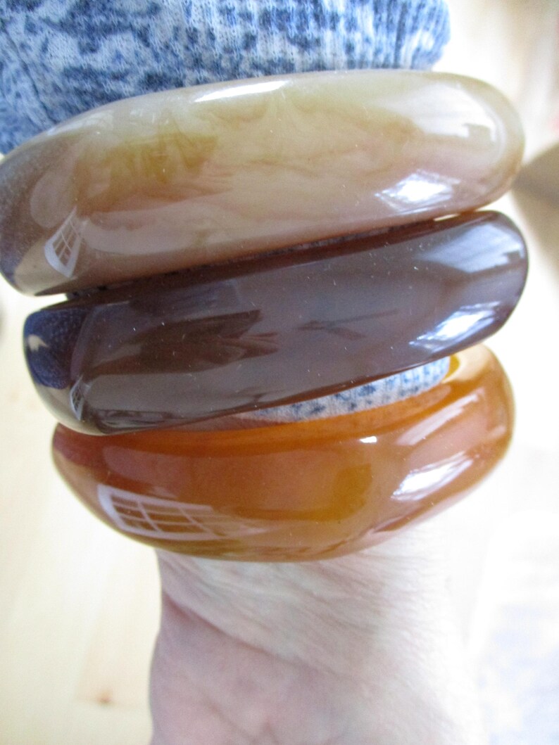 K&ouml;nnte beinhalten: Drei gl&auml;nzende, ovale Armreifen in Braun-, Bernstein- und Karamellt&ouml;nen. Die Armb&auml;nder sind &uuml;bereinander gestapelt und zeigen ihre glatten, polierten Oberfl&auml;chen. Die Armreifen scheinen aus einem durchscheinenden Material zu bestehen.