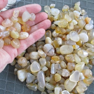May include: A collection of polished, tumbled gemstones in shades of yellow, cream, and amber. Some stones are held in a hand, while the rest are scattered on a surface. The stones vary in size and shape, with a smooth, glossy finish.