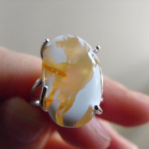 May include: A silver ring featuring a large, oval-shaped gemstone. The stone has a white base with swirling patterns of orange and yellow. The ring's setting has four prongs securing the stone.