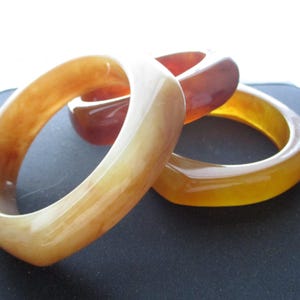 K&ouml;nnte beinhalten: Drei quadratisch geformte Armreifen in den Farbt&ouml;nen Bernstein, Braun und Gelb. Die durchscheinenden Armb&auml;nder sind auf einer dunklen Oberfl&auml;che gestapelt und zeigen ihre glatte, polierte Oberfl&auml;che. Das Licht f&auml;ngt die Kurven des Schmucks ein.