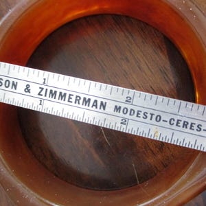 K&ouml;nnte beinhalten: Ein vintage, bernsteinfarbenes, quadratisch geformtes Armreif-Armband. Ein Lineal liegt &uuml;ber dem Armband und misst etwa 7,6 cm im Durchmesser. Auf dem Lineal steht "BENSON & ZIMMERMAN MODESTO-CERES-NEW".