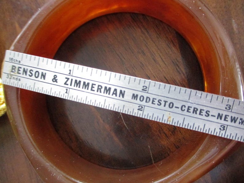 K&ouml;nnte beinhalten: Ein vintage, bernsteinfarbenes, quadratisch geformtes Armreif-Armband. Ein Lineal liegt &uuml;ber dem Armband und misst etwa 7,6 cm im Durchmesser. Auf dem Lineal steht "BENSON & ZIMMERMAN MODESTO-CERES-NEW".