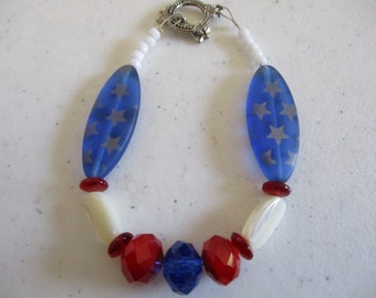 Könnte beinhalten: Ein Perlenarmband mit zwei länglichen blauen Glasperlen mit silbernen Sternmustern, weißen Saatperlen, glatten weißen ovalen Perlen sowie facettierten roten und blauen Kristallperlen, abgeschlossen mit einem silberfarbenen Knebelverschluss.