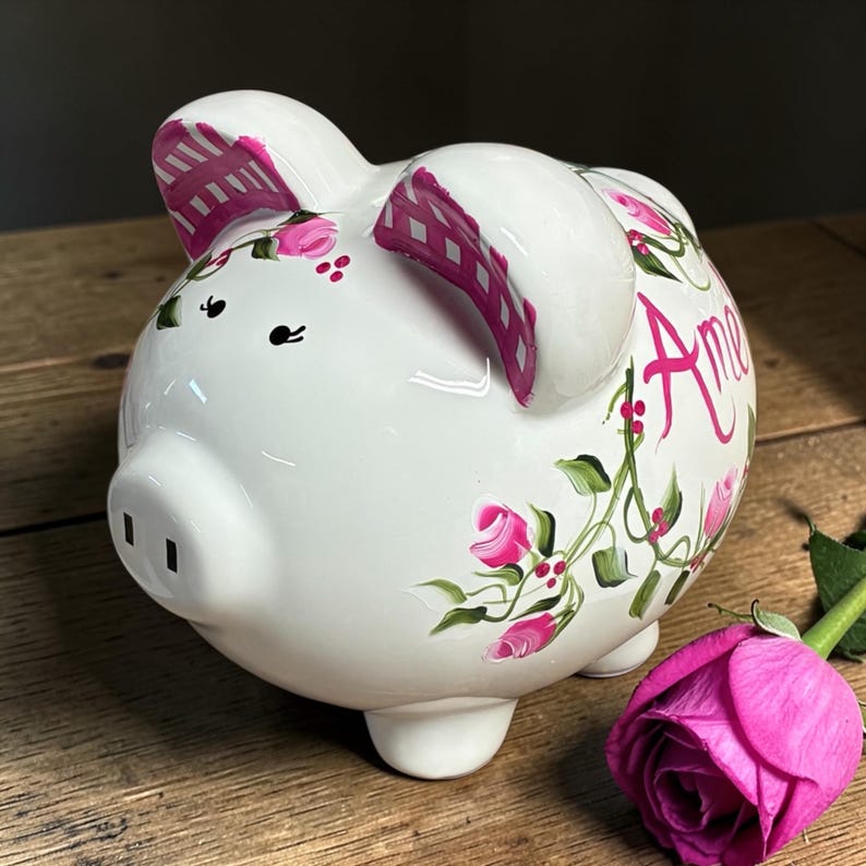 May include: A white ceramic piggy bank featuring hand-painted pink roses with green leaves and vines. The ears are accented with a pink checkered pattern. The name "Amelia" is written in pink script on the side of the pig, which has black eyes.