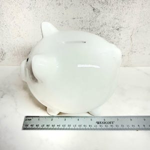 May include: A glossy white ceramic piggy bank with a coin slot on top, positioned behind a metal Westcott ruler that shows the object is approximately 6 inches long. The piggy bank features rounded ears and a snout, with a smooth, reflective surface.