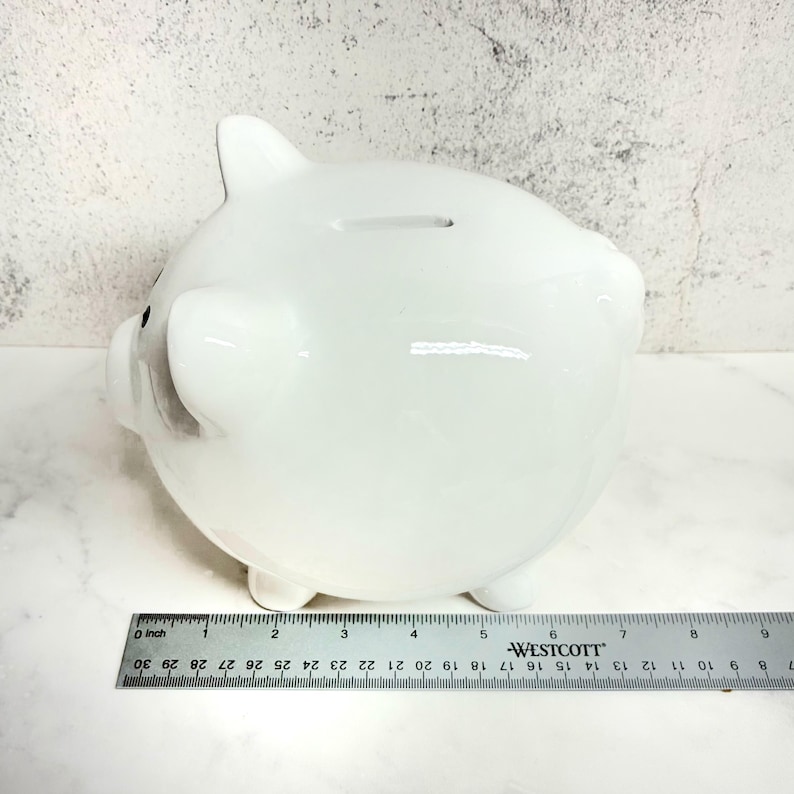 May include: A glossy white ceramic piggy bank with a coin slot on top, positioned behind a metal Westcott ruler that shows the object is approximately 6 inches long. The piggy bank features rounded ears and a snout, with a smooth, reflective surface.