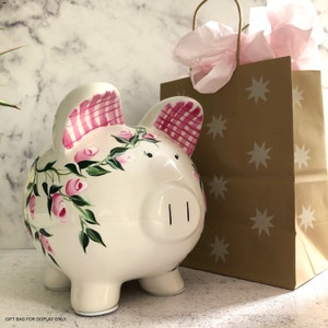 May include: A white ceramic piggy bank featuring hand-painted pink rose vines and green leaves across the body. The ears are decorated with a pink and white gingham check pattern. A brown paper gift bag with white star prints is positioned behind.