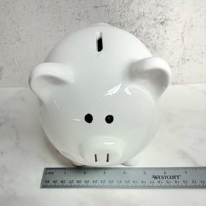 May include: A glossy white ceramic piggy bank with two black painted eyes and two black vertical lines for nostrils. A coin slot is located on the top of the head. A metal Westcott ruler is positioned at the base, showing a width of 7 inches.