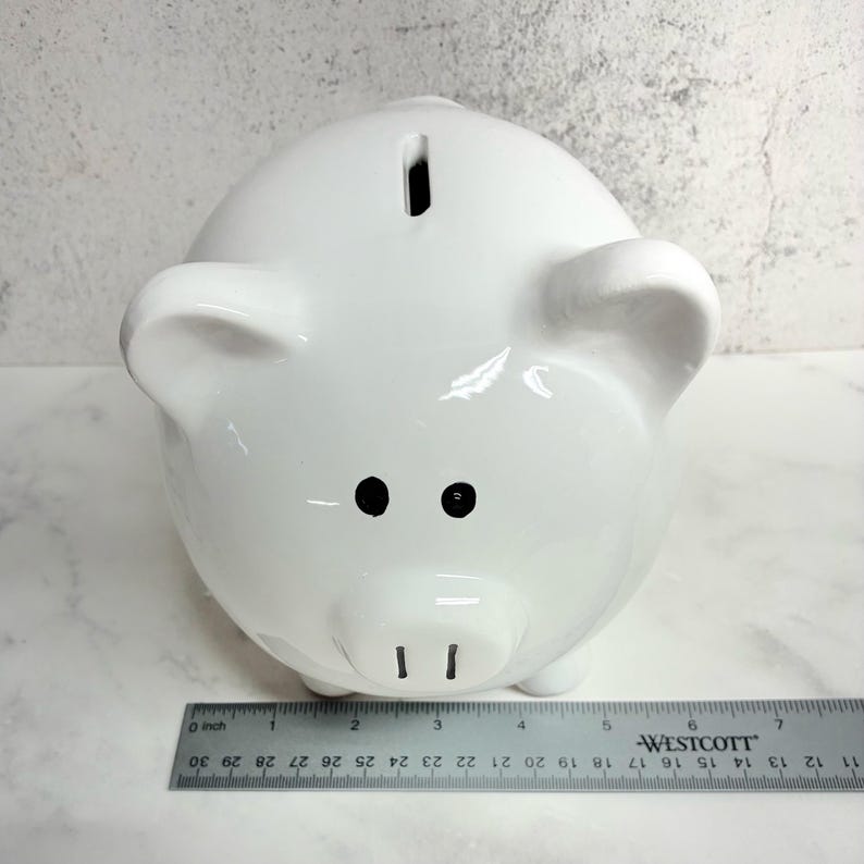 May include: A glossy white ceramic piggy bank with two black painted eyes and two black vertical lines for nostrils. A coin slot is located on the top of the head. A metal Westcott ruler is positioned at the base, showing a width of 7 inches.