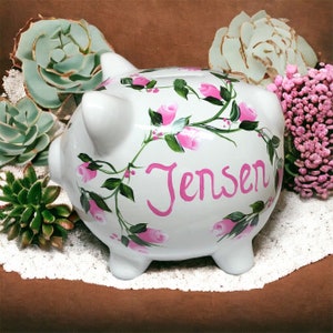 May include: A white ceramic piggy bank featuring a hand-painted design of pink rosebuds and green vines wrapping around the body. The name Jensen is written in large, bright pink cursive script across the center of the piggy bank's side.