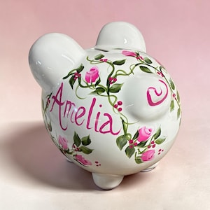 May include: A white ceramic piggy bank featuring a hand-painted design of green vines, small red berries, and pink rose buds. The name Amelia is written in a cursive pink script on the side of the piggy bank, which has two ears and a tail.