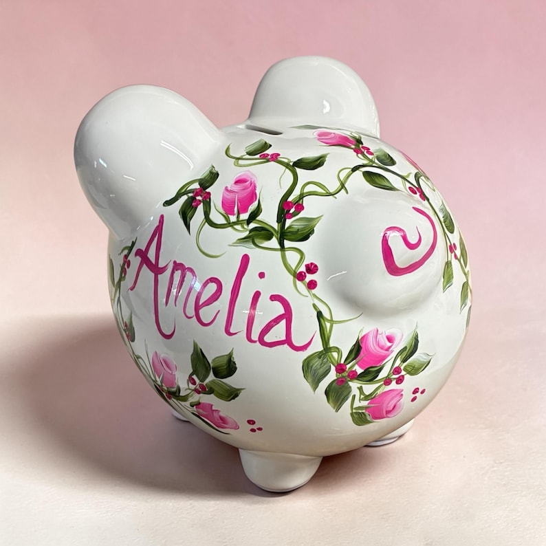 May include: A white ceramic piggy bank featuring a hand-painted design of green vines, small red berries, and pink rose buds. The name Amelia is written in a cursive pink script on the side of the piggy bank, which has two ears and a tail.