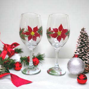 May include: Two clear glass wine glasses featuring hand-painted red poinsettia flowers with yellow centers and green pine needle accents. The base of each glass is decorated with small red berries and green pine needles, creating a festive look.