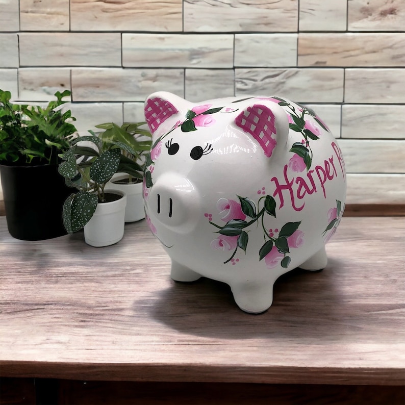 May include: A white ceramic piggy bank featuring hand-painted pink roses with green leaves and a pink checkered pattern on the ears. The name Harper R is written in pink script on the side. The piggy bank has black painted eyes with eyelashes.