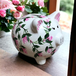 May include: A white ceramic piggy bank featuring a hand-painted floral design of pink roses with green leaves and vines. The pig has pink striped ears, black painted eyes, and a coin slot on its back. The surface is glossy and smooth.